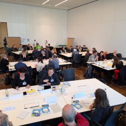 Auf dem Foto sind Menschen zu sehen, die an Gruppentischen sitzen. Sie sind miteinander im Gespräch und schreiben auf Moderationskarten.