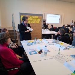 Auf dem Bild sind Menschen zu sehen, die an einem Gruppentisch sitzen. Eine Person steht vorne an einer Pinnwand und pinnt Moderationskarten an.